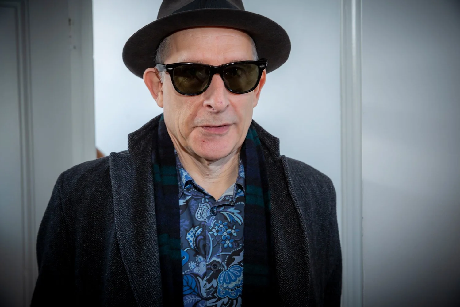 A man wearing a black fedora, dark sunglasses, a gray coat, a blue patterned shirt, and a scarf, standing indoors.