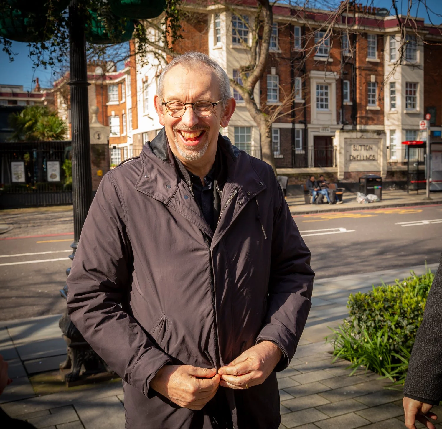 A middle-aged man with glasses and a gray beard is smiling and appears to be laughing. He is wearing a dark gray jacket and standing outdoors on a sidewalk, with a row of modern brick buildings and trees in the background.