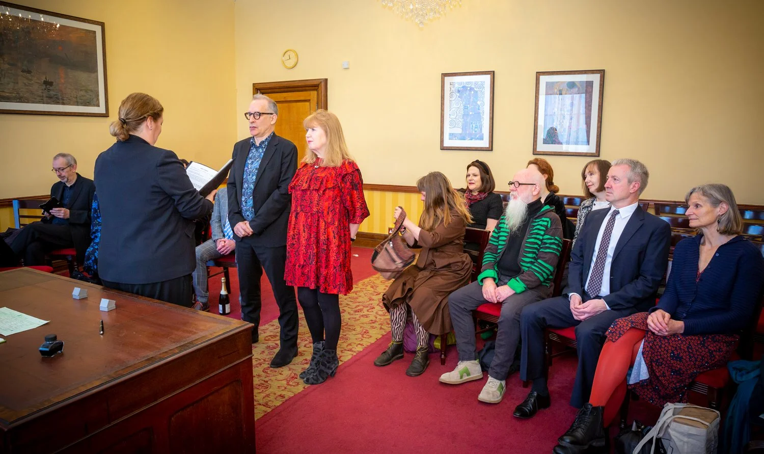People at a formal event with some standing and some seated, inside a room with yellow walls: a woman reads from a folder, two men with glasses and a woman in a red dress stand in front of her, and others sit observing.