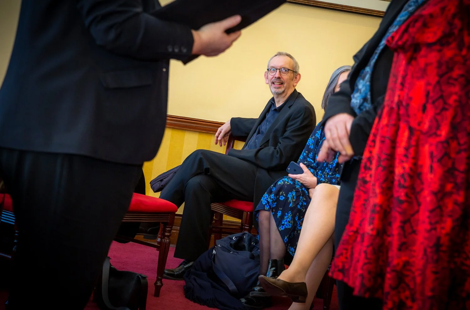 A man with glasses and a beard in a black suit sitting on a red cushioned chair, smiling and talking to a woman in a blue floral dress. They are in a room with yellow walls and other people standing or sitting nearby.