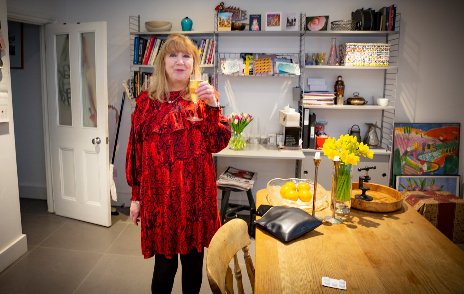 Woman in red dress holding a glass of wine in a home dining room with flowers and artwork.