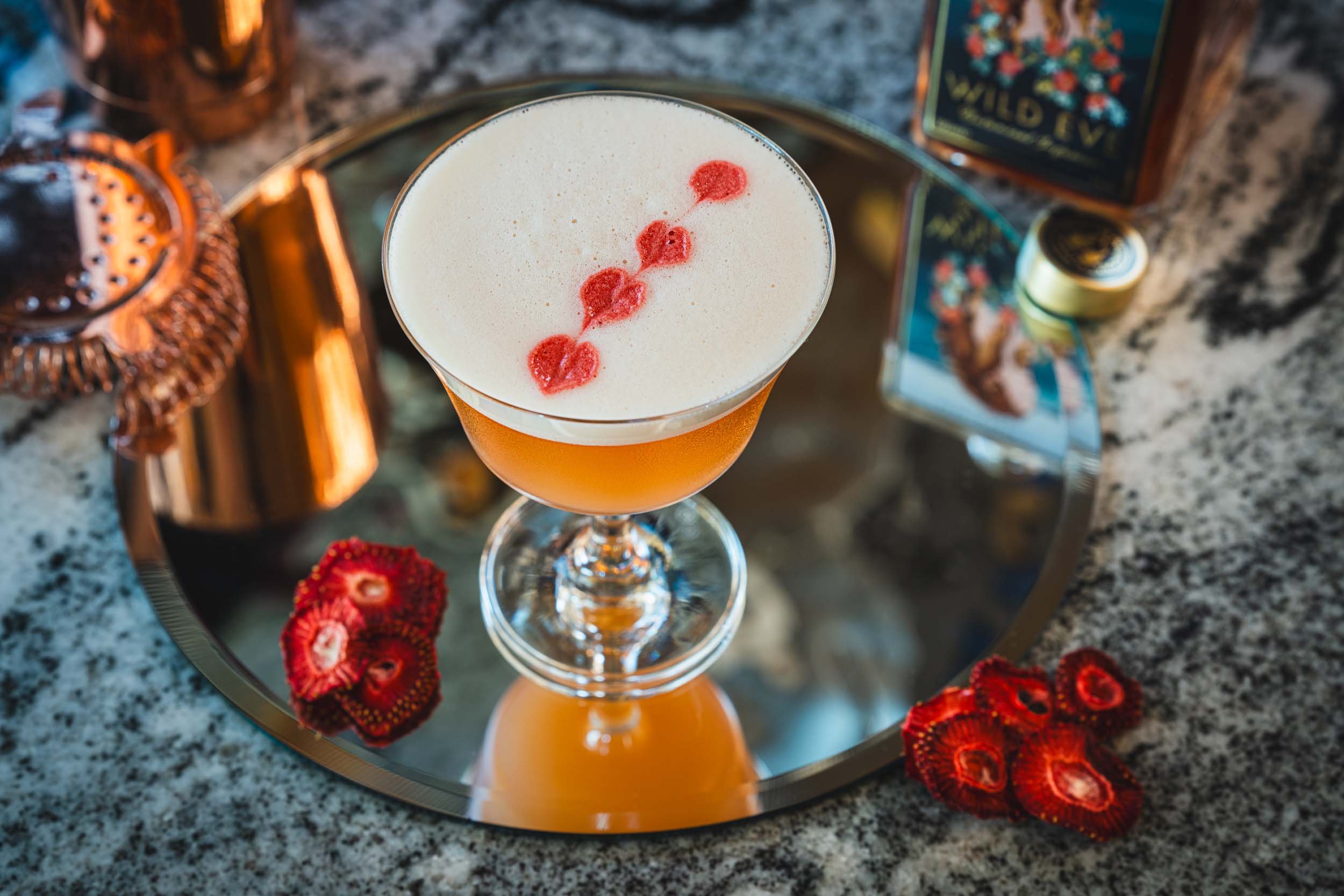 A cocktail with a frothy white top garnished with red fruit, served in a coupe glass on a reflective silver tray, with dried strawberries, a copper drinkmixer, and a bottle of Wild Eyed whiskey in the background.