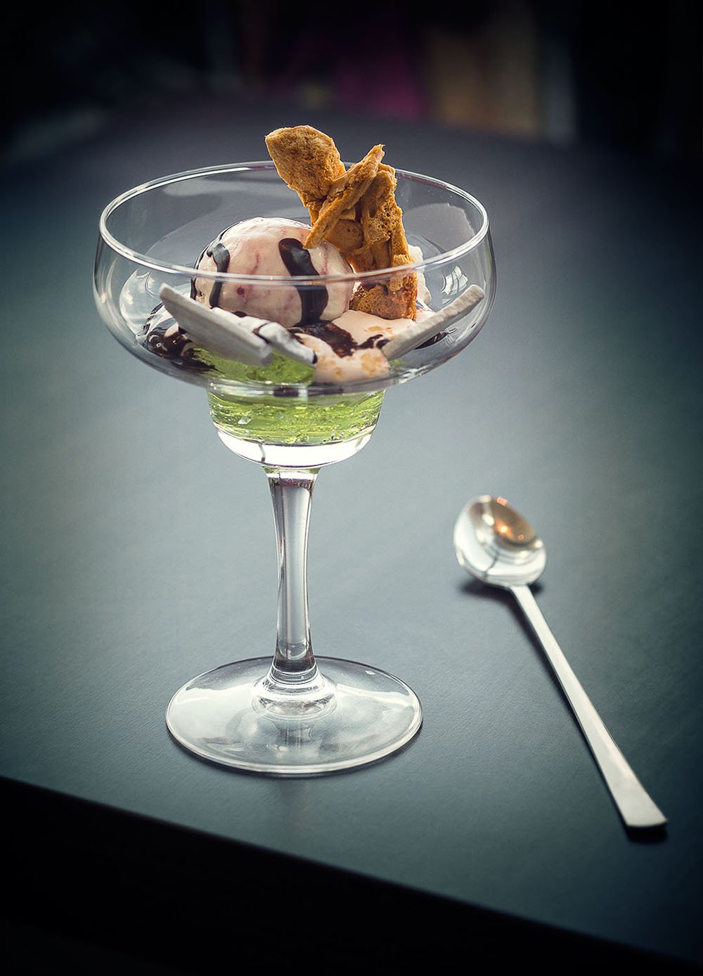 A dessert in a glass coupe with ice cream, cookie pieces, and chocolate syrup, accompanied by a silver spoon on a dark table.