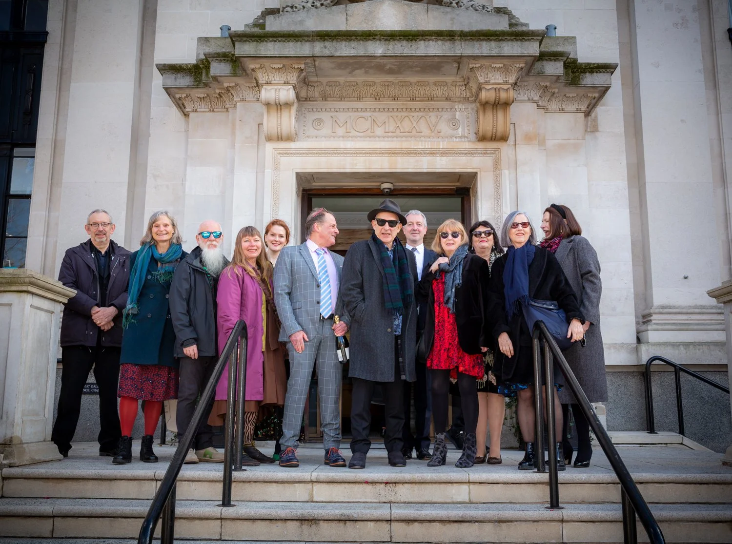 Group of people standing on the steps outside a government building, dressed in formal and semi-formal attire, with some wearing sunglasses and holding a wine bottle, during the daytime.