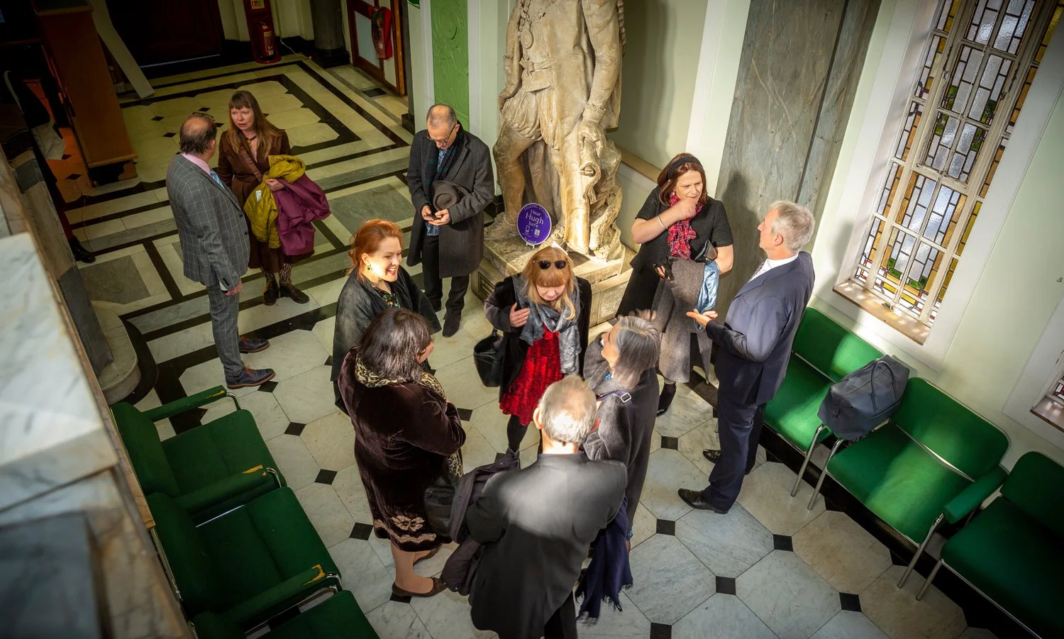 Group of people having a conversation in a hallway with polished floors, green chairs, and stained glass windows at an art gallery or museum.