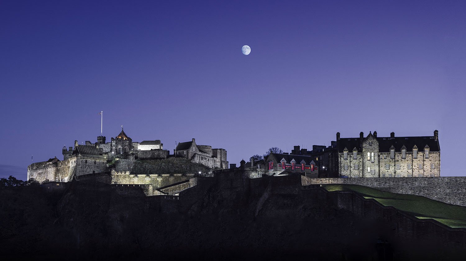 Nighttime view of  historic Edinburgh Castle with stone walls and turrets under a clear sky with the moon.