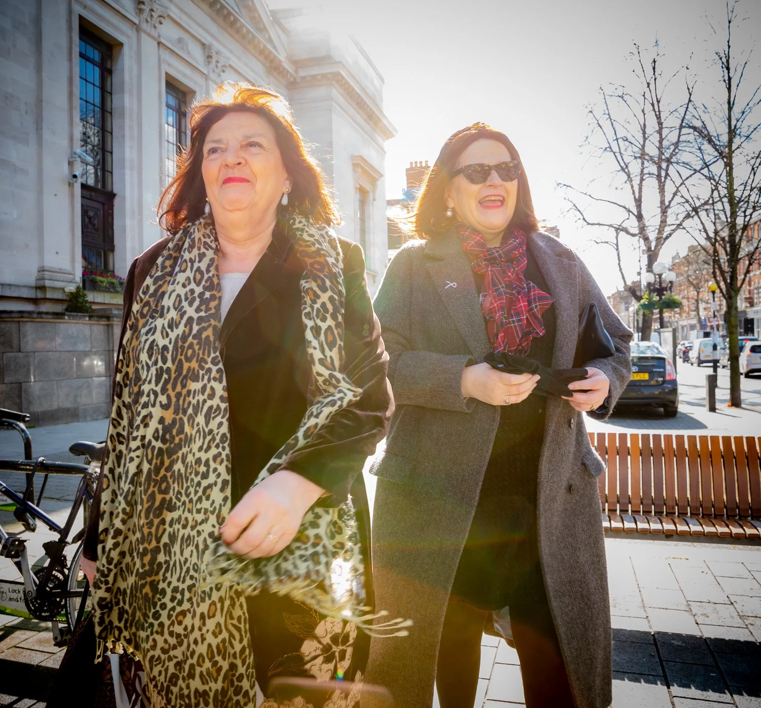 Two women walking outdoors on a sunny day, one wearing a leopard print scarf and coat, the other wearing sunglasses and a grey coat, with trees and a building in the background.