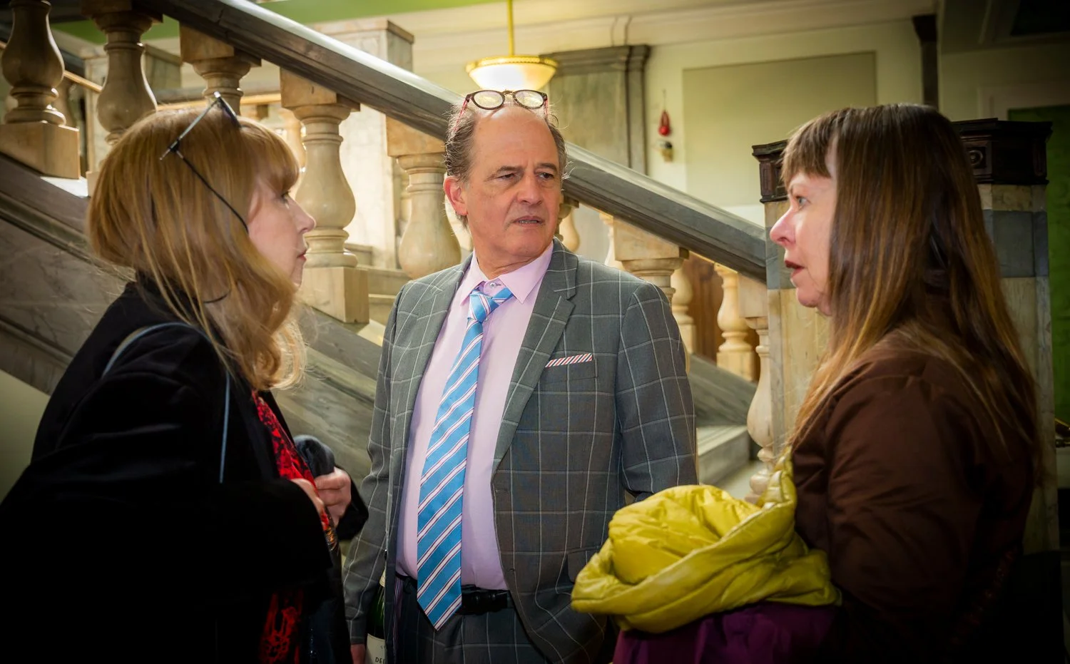 Three people having a conversation in a building with a staircase in the background. A man in a plaid suit and women in jackets are talking.