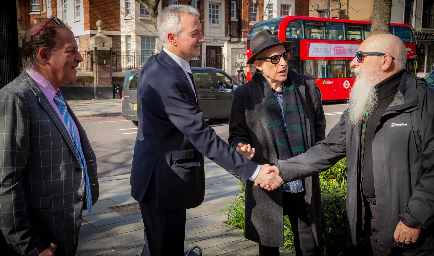 A group of four men meeting and shaking hands on a city sidewalk. One man is wearing a suit, two are wearing dark coats and hats, and one has a long white beard and glasses.