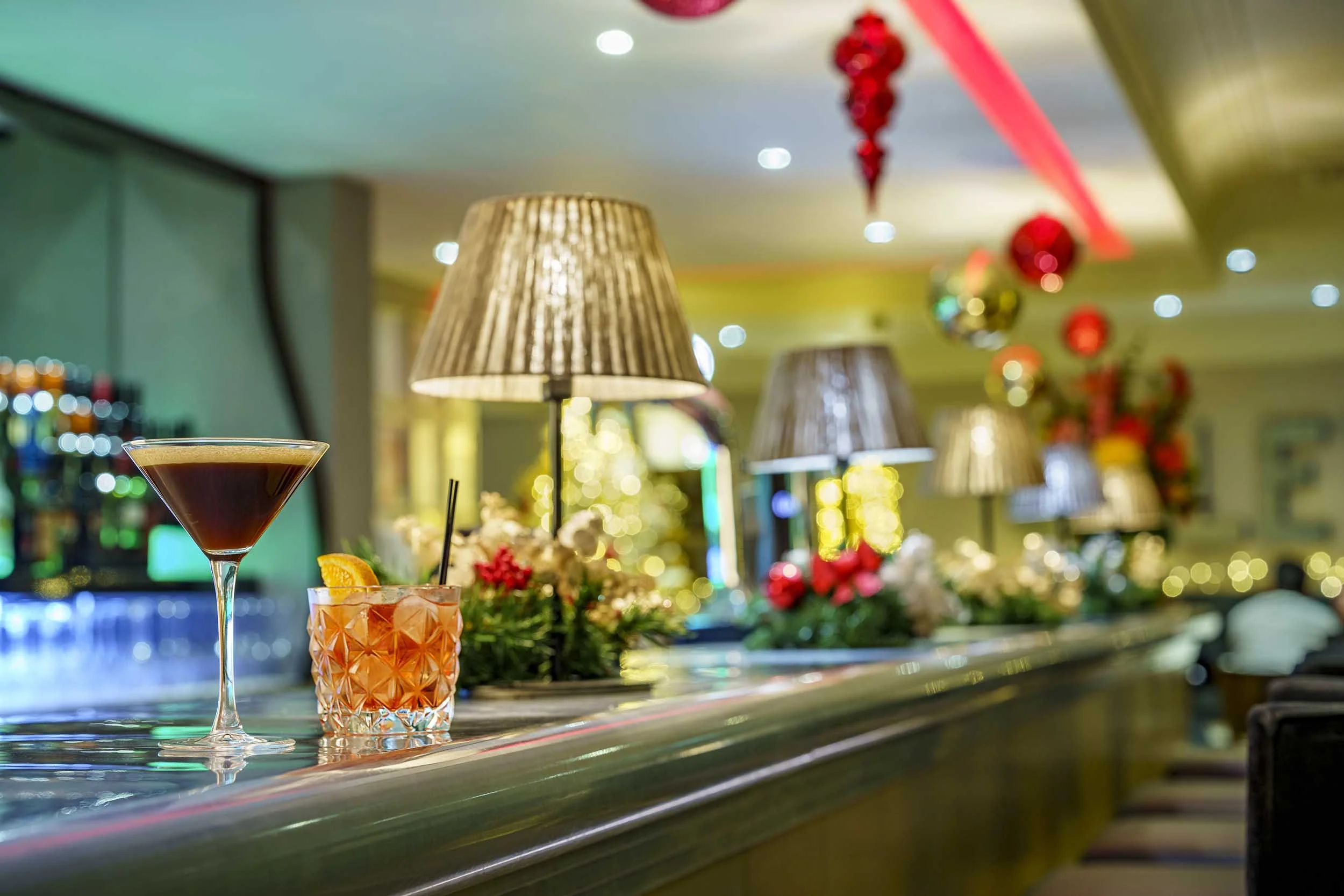 Close-up view of two cocktails on a bar counter with festive Christmas decorations and lamps in the background, likely in a restaurant or bar set for a celebration.