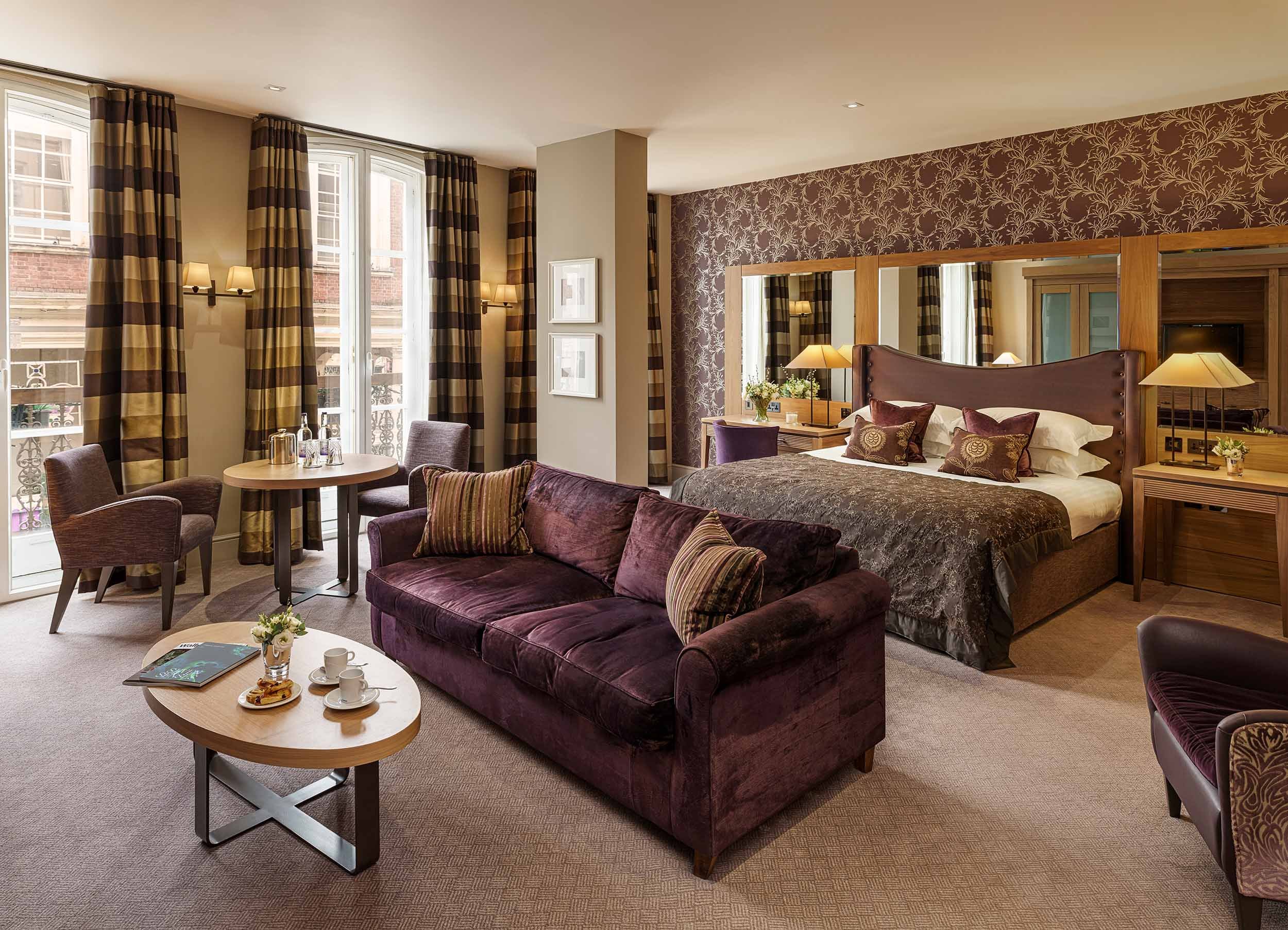 A luxurious hotel suite featuring a bedroom with a large bed, purple headboard, and multiple pillows. There is a sitting area with a purple velvet sofa, a small round table with tea set, and a round dining table with chairs near windows with striped 