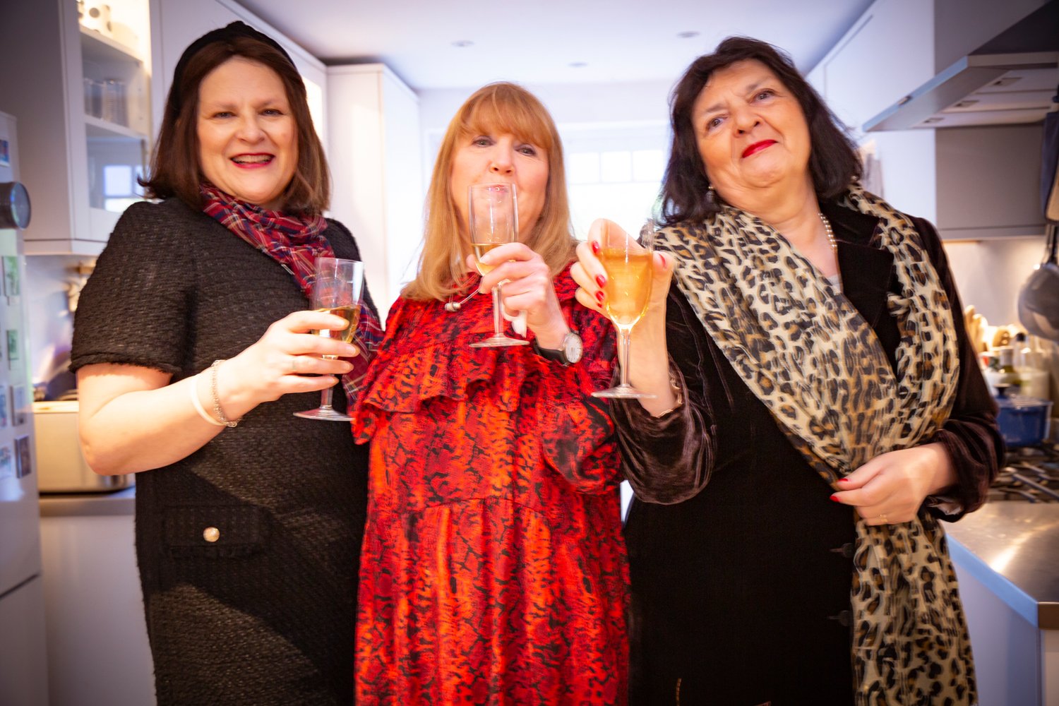 Three women celebrating with drinks in a kitchen, smiling and making faces.