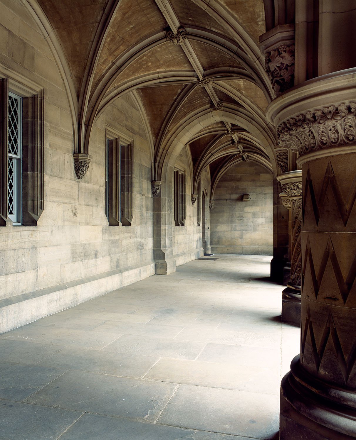 A stone Cloistered walkway or corridor with pointed Gothic arches, small windows with diamond-shaped panes, and ornate columns with carved capitals.
