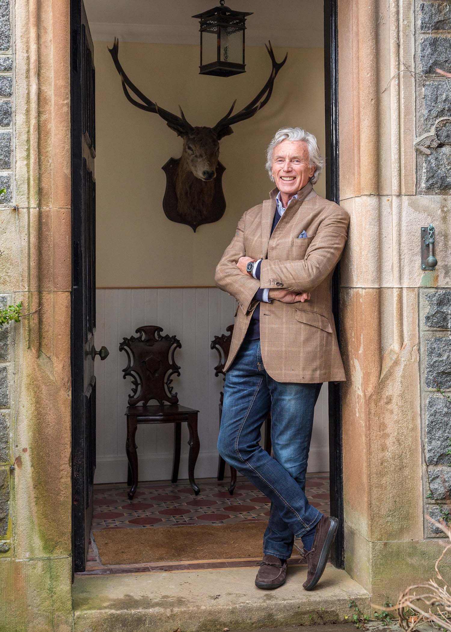 A smiling man with gray hair wearing a brown blazer and jeans, standing at the doorway of a house. Inside, there is a mounted deer head on the wall and two ornate chairs.