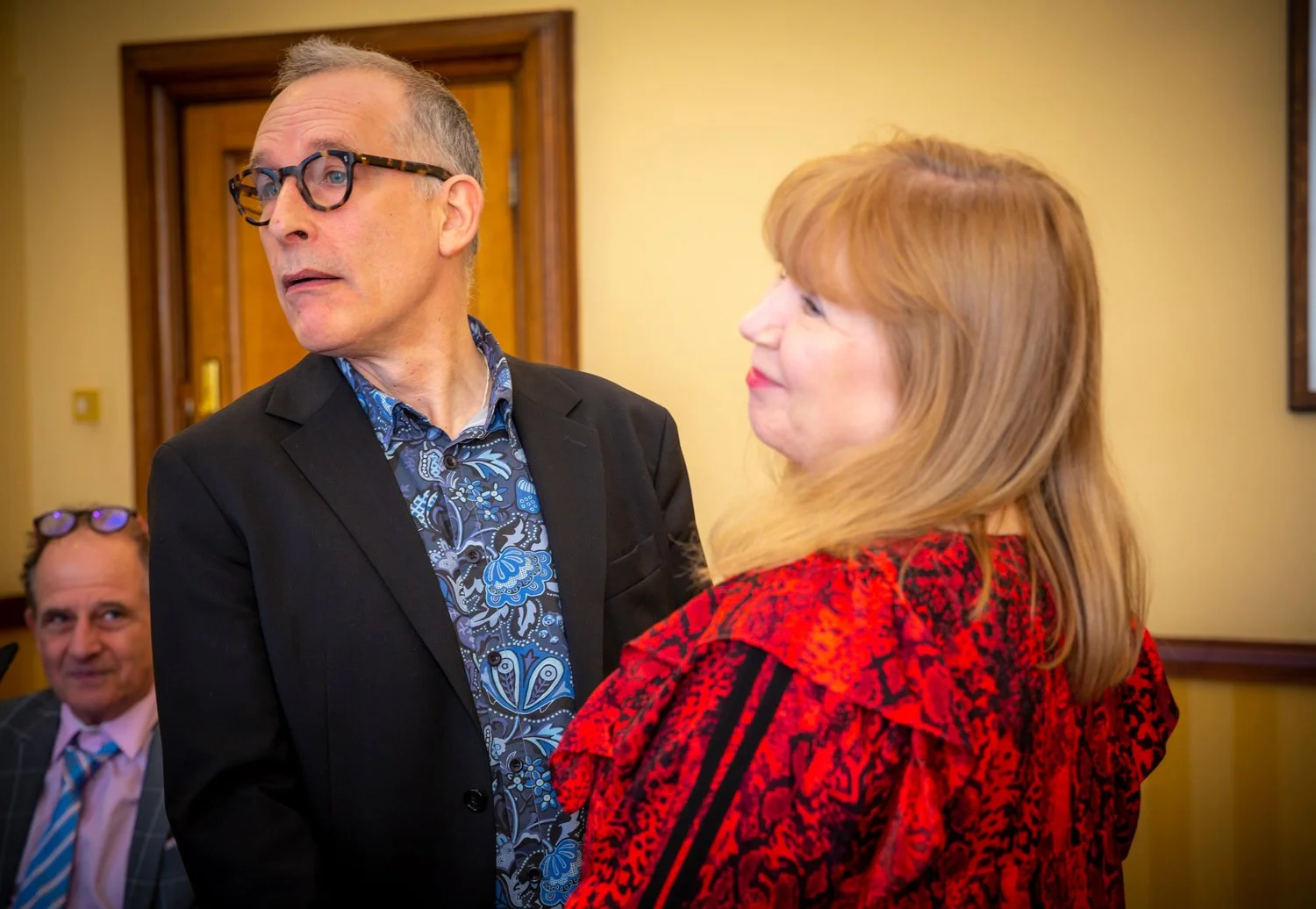 A man with glasses and a dark blazer looking to the side, a woman with blonde hair in a red and black patterned dress facing him, and another man in a suit and glasses in the background.