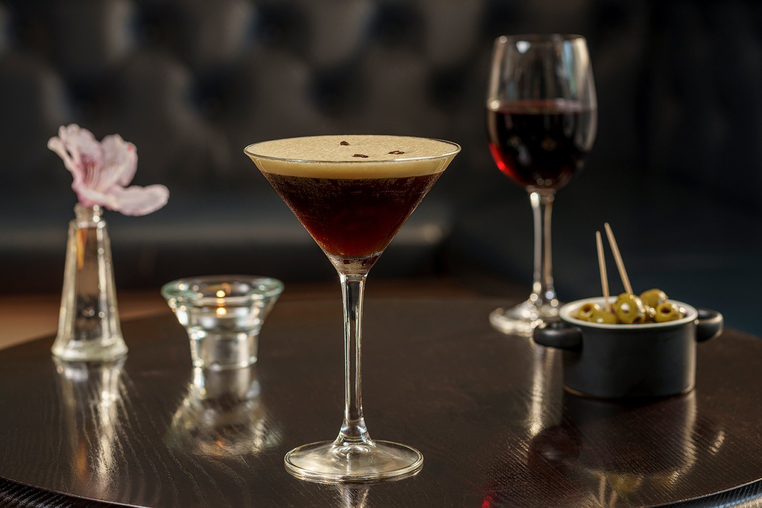 A dark Espresso Martini cocktail with a foam top and coffee bean garnish in a martini glass, a glass of red wine, a small bowl of green olives with toothpicks, and a small vase with a pink flower on a dark table.