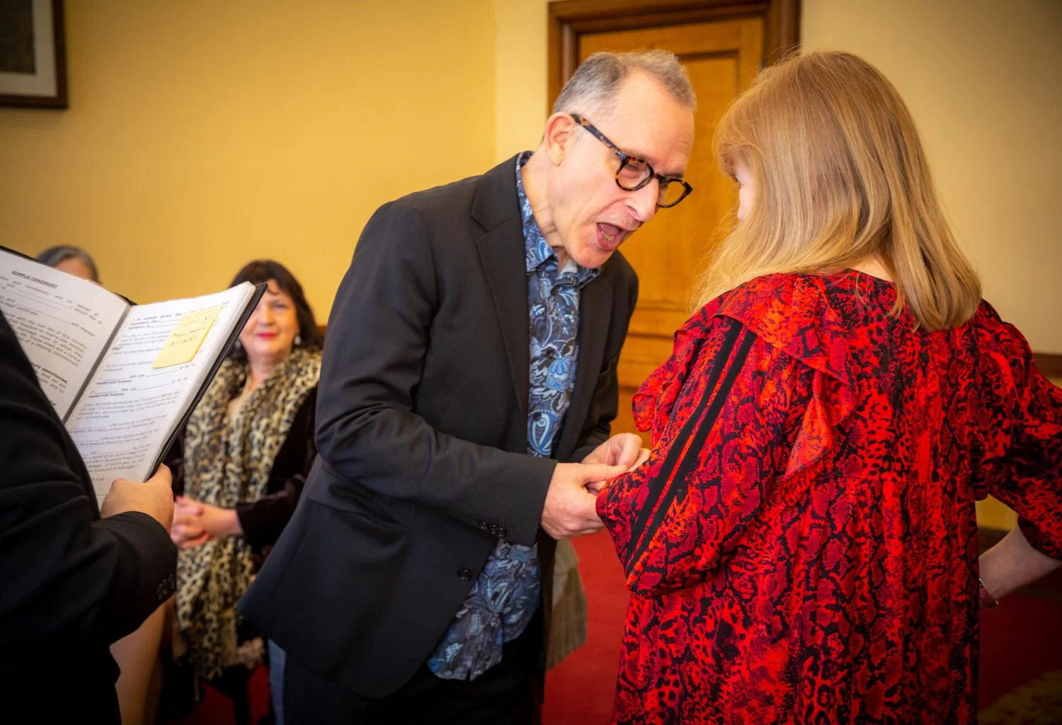 A man with glasses in a suit holding hands with a woman in a red patterned dress at a formal event, with other people seated in the background.