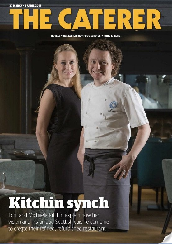 Tom and Michaela Kitchin stand inside a restaurant, smiling at the camera. Tom is wearing a chef's jacket, and Michaela is dressed in black. The background shows a modern dining area with tables and chairs. The magazine cover headline reads 'The Cate