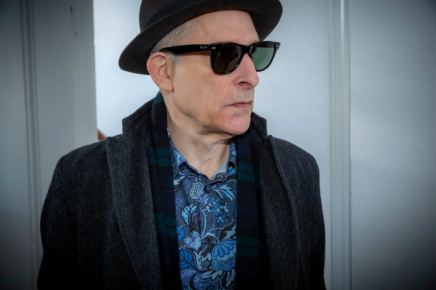 A man wearing a black fedora, black sunglasses, a patterned blue shirt, and a dark textured coat, standing indoors.