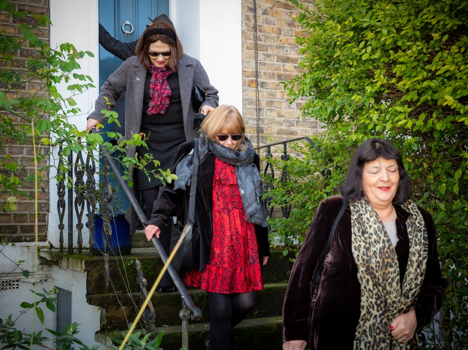 Three women, dressed stylishly, descend outdoor stone stairs near a brick house door with a blue frame, surrounded by greenery.
