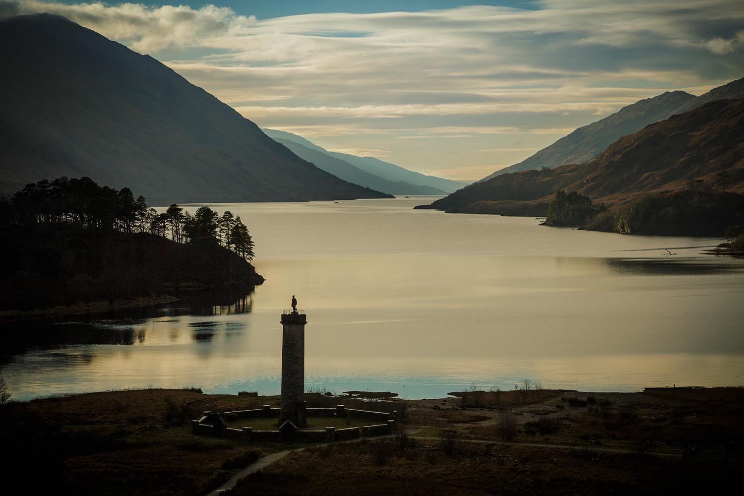 A scenic landscape of Loch Shiel, a large body of water surrounded by mountains with clouds in the sky. In the foreground, there is the tall Glenfinnan Monument with a person standing on top, and some trees and pathways around it.