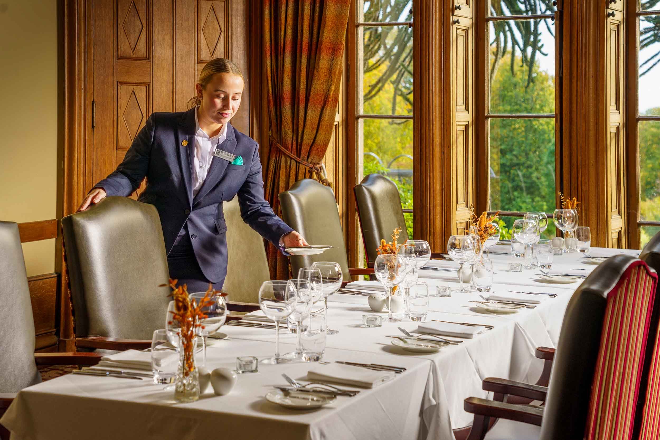 A woman in a formal navy blazer is setting a dining table in a bright, wood-paneled room with large windows and autumnal decor.