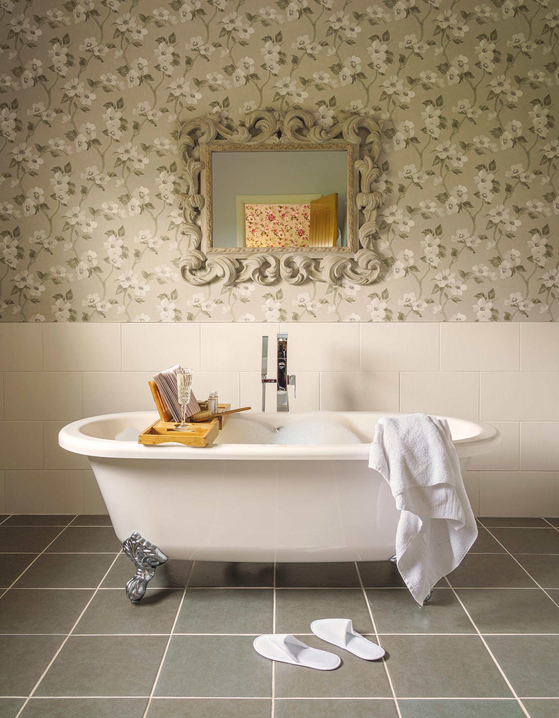 A vintage clawfoot bathtub with a wooden bath tray holding a glass of champagne, bath brush, and small bottles, with a white towel hanging over the side, in front of a floral patterned wallpapered wall with a decorative mirror, on a checked tile floo