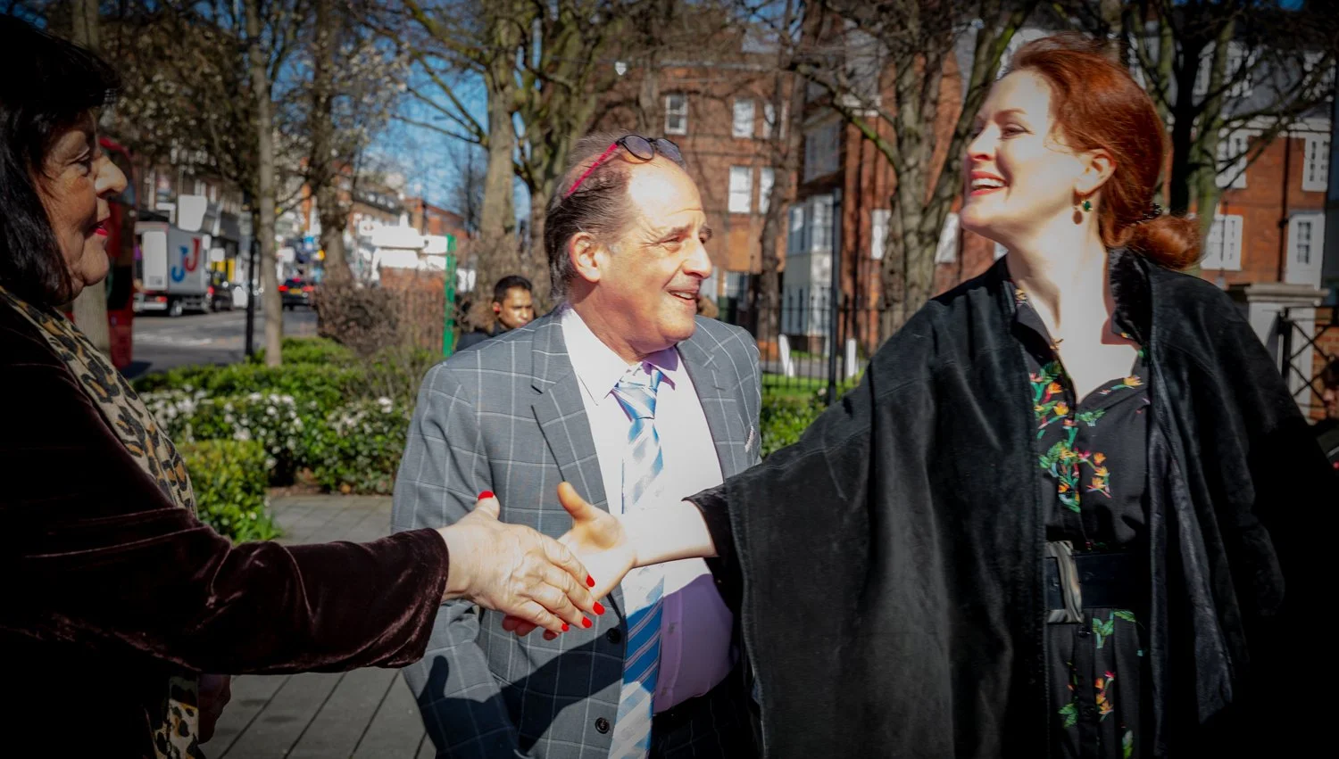 Three women greeting each other with a handshake outdoors in a park on a sunny day. The women are smiling and dressed in casual and semi-formal clothing.