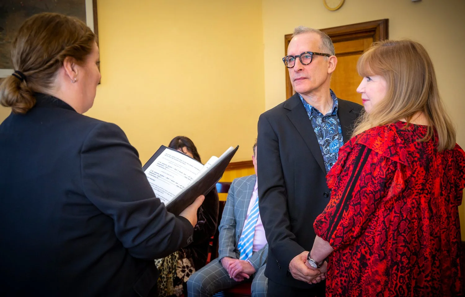 A woman in a black suit reads from a paper while two people, a man and a woman, hold hands and listen at a formal event in a room with yellow walls.
