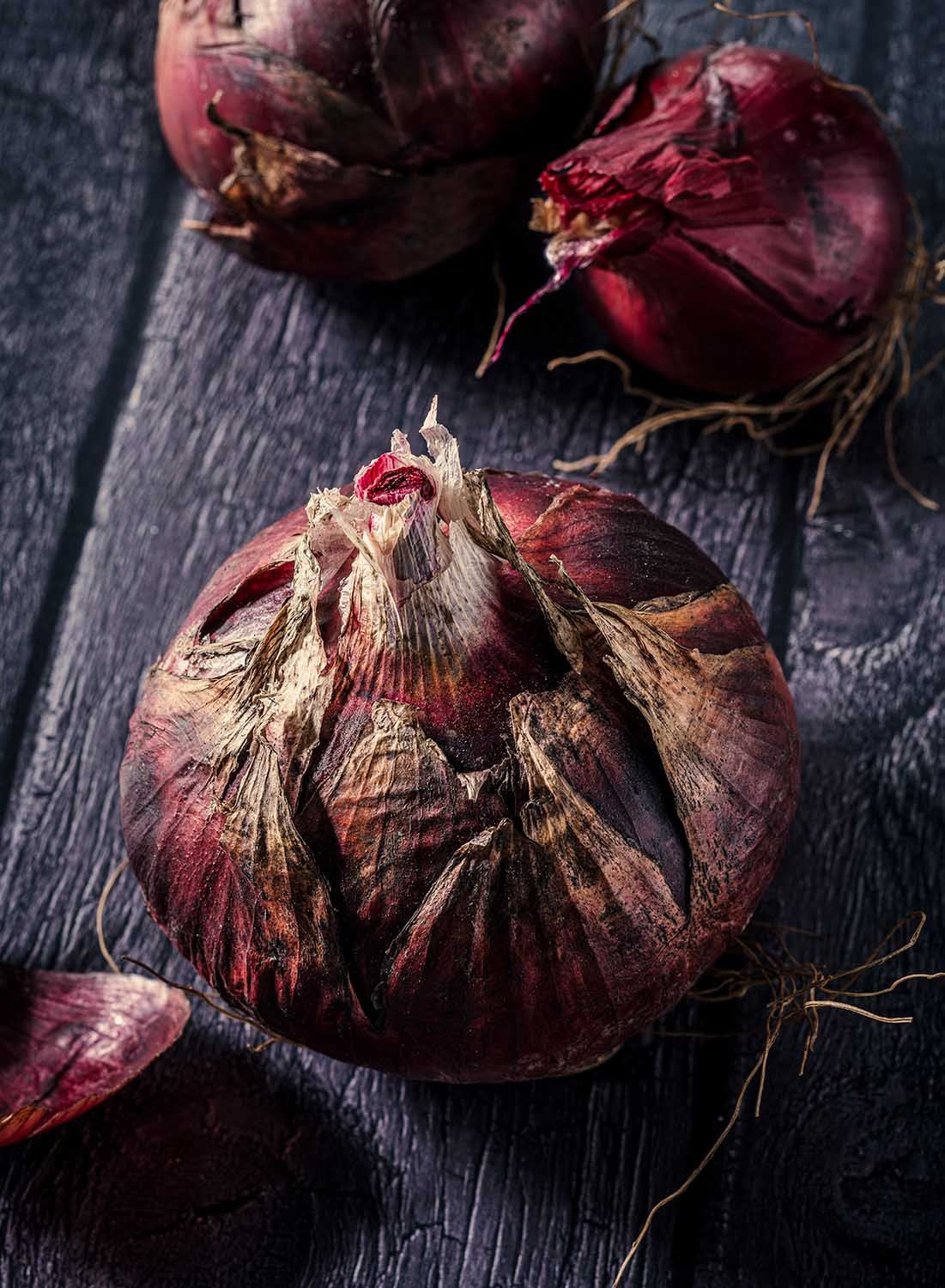 A close-up of three dried red onions with papery skin and roots, placed on a dark wooden surface.