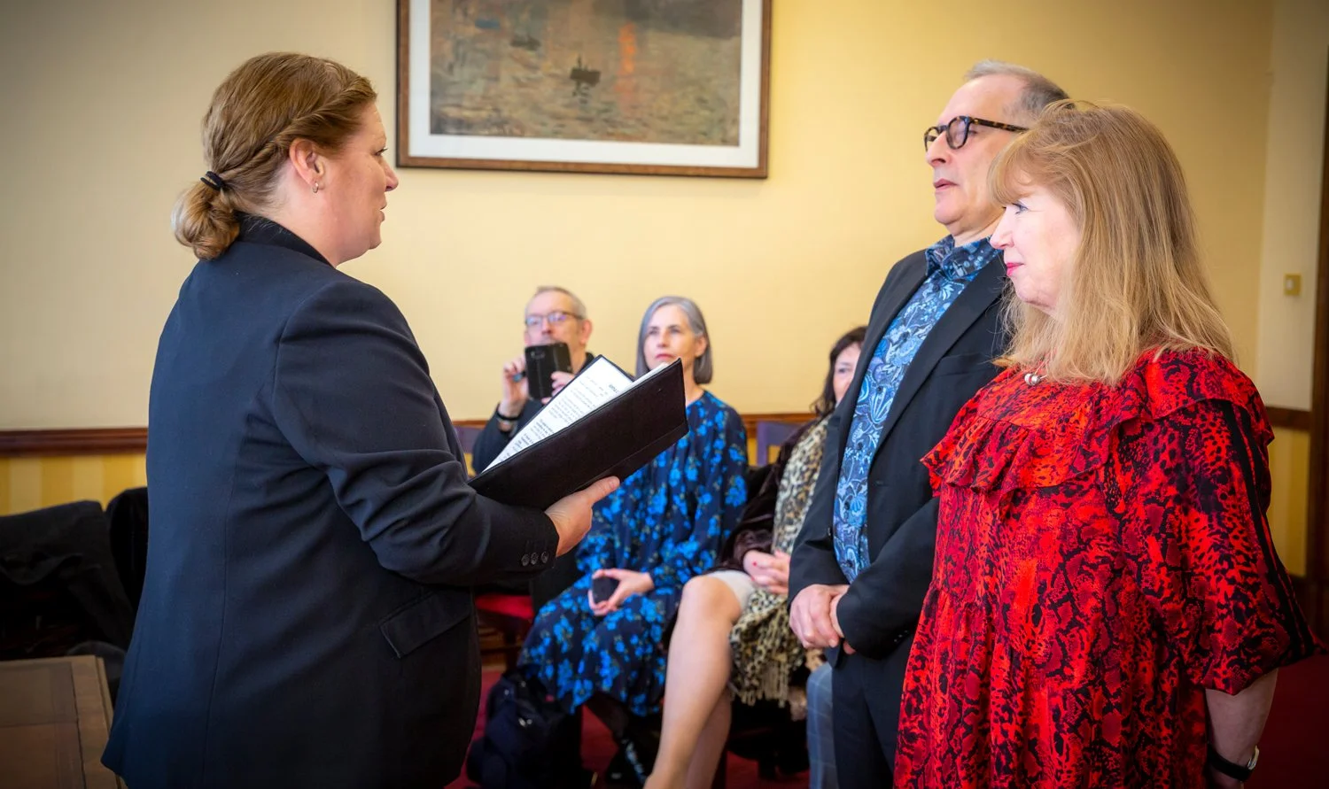 A woman in a black blazer reads from a folder in front of a small group of people during a formal event in a room with yellow walls and a framed painting.