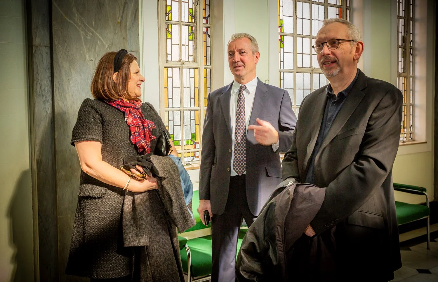 Three adults having a conversation inside a building with stained glass windows in the background. The woman on the left is smiling and holding a bag, the man in the middle is gesturing with his finger, and the man on the right is listening with a co