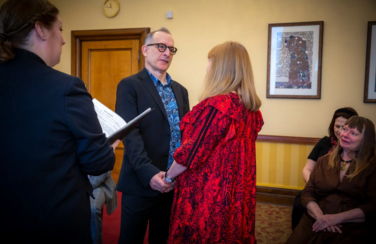 A woman in a red patterned dress holding hands with a man in a black suit and glasses, facing each other in a room with art on the walls, while three other women are seated nearby, observing the interaction.