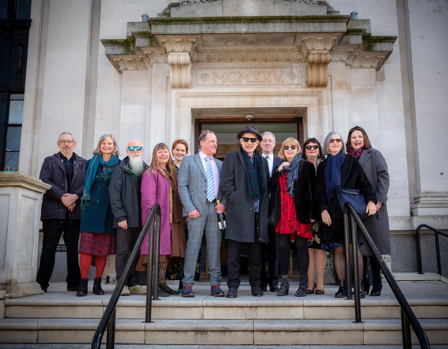 A group of people standing on steps outside a historical building with stone architecture, posing for a photo. Some are dressed in formal attire, while others are in casual clothing. The building has an inscription with Roman numerals above the doorw