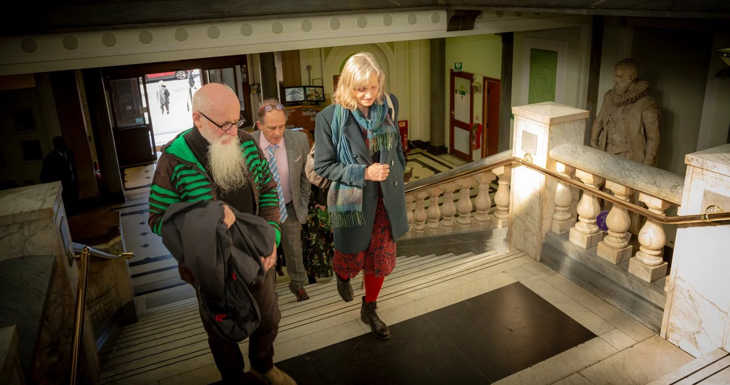 People walking up a staircase inside a building, with a large statue of a man in historical clothing at the top of the stairs.