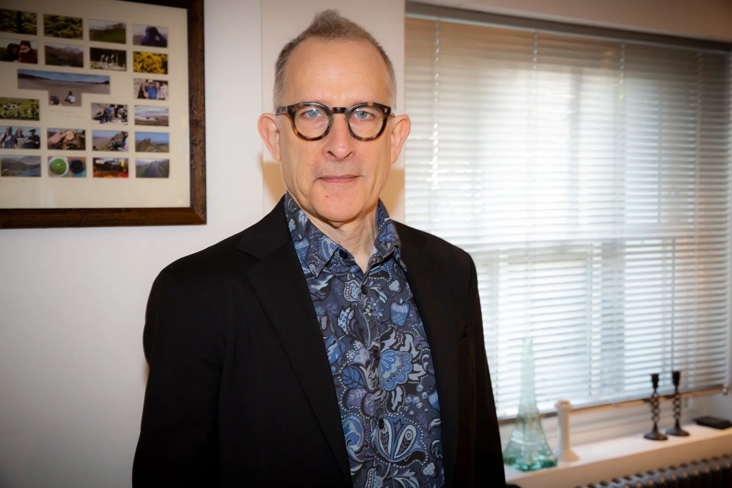 A middle-aged man with glasses and short hair wearing a dark blazer and a blue patterned shirt, standing indoors near a window with blinds, with framed photos on the wall behind him.
