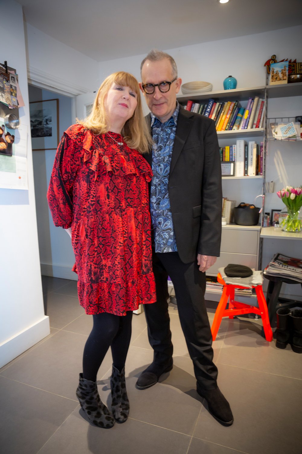 A woman with red hair wearing a red patterned dress and leopard print boots standing next to a man with glasses wearing a black suit and a blue patterned shirt, indoors in a room with shelves of books, a small table with a vase of pink tulips, and a 