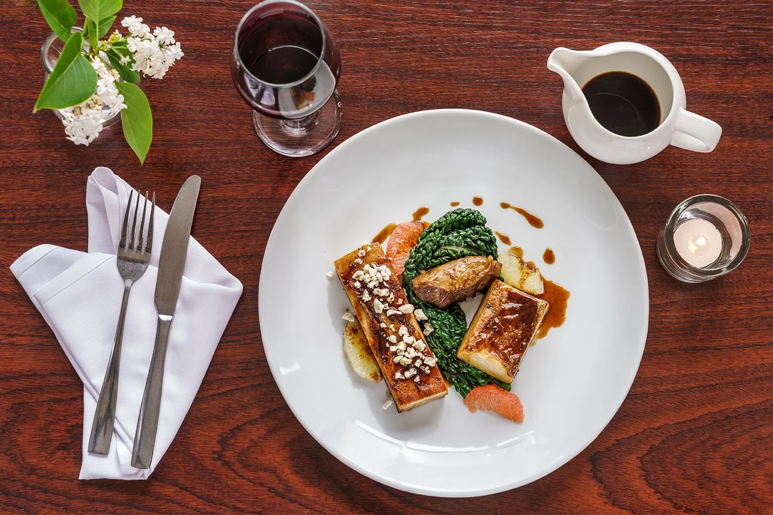 A top-down view of a dinner table with a white plate of gourmet food, a glass of red wine, a cup of black coffee, a small bowl of flowers, a folded napkin with a fork and knife, and a lit candle.