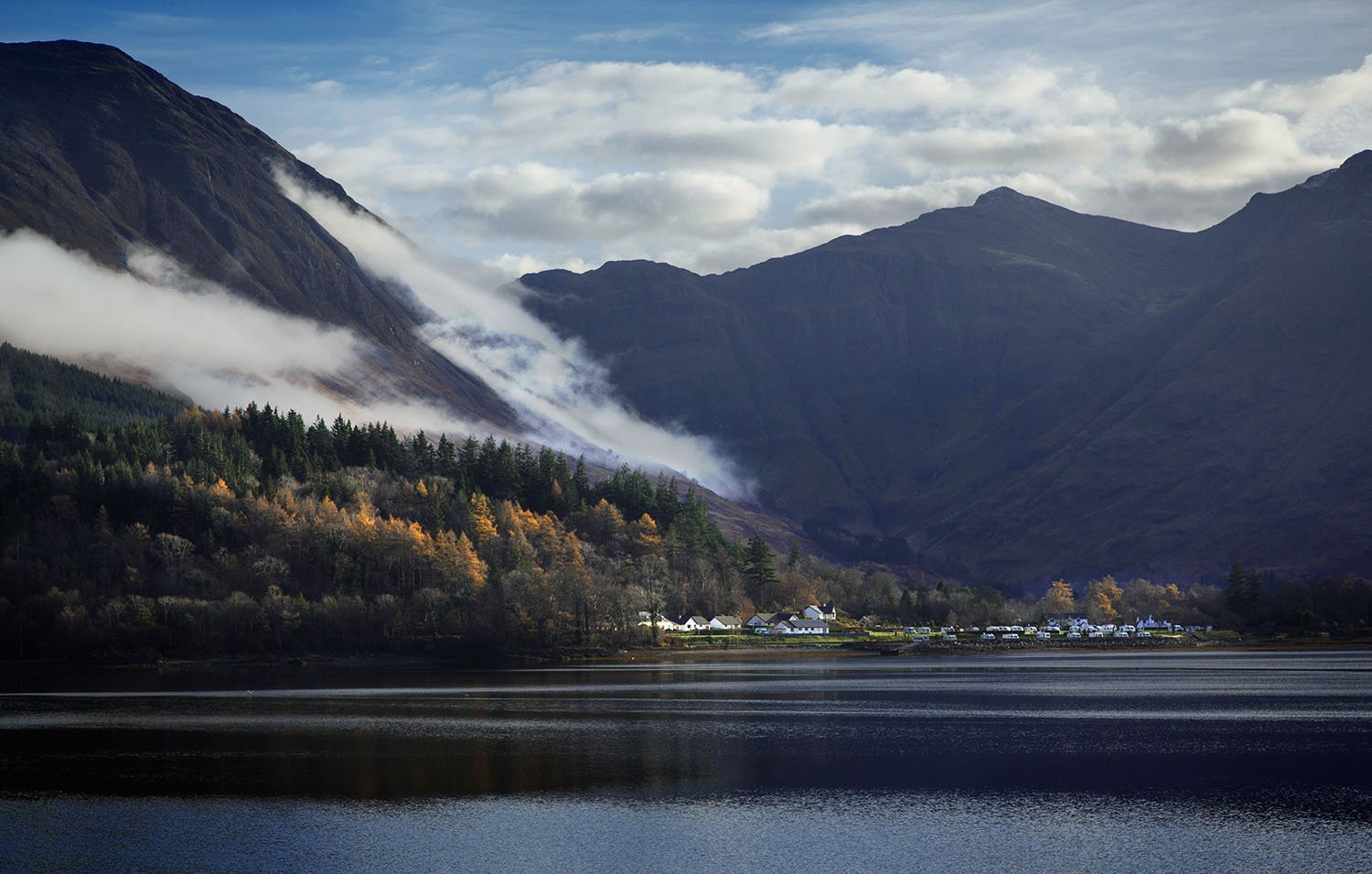 Scenic view of a lake with mountains in the background, trees along the shoreline, and Glen Coe village with white buildings.