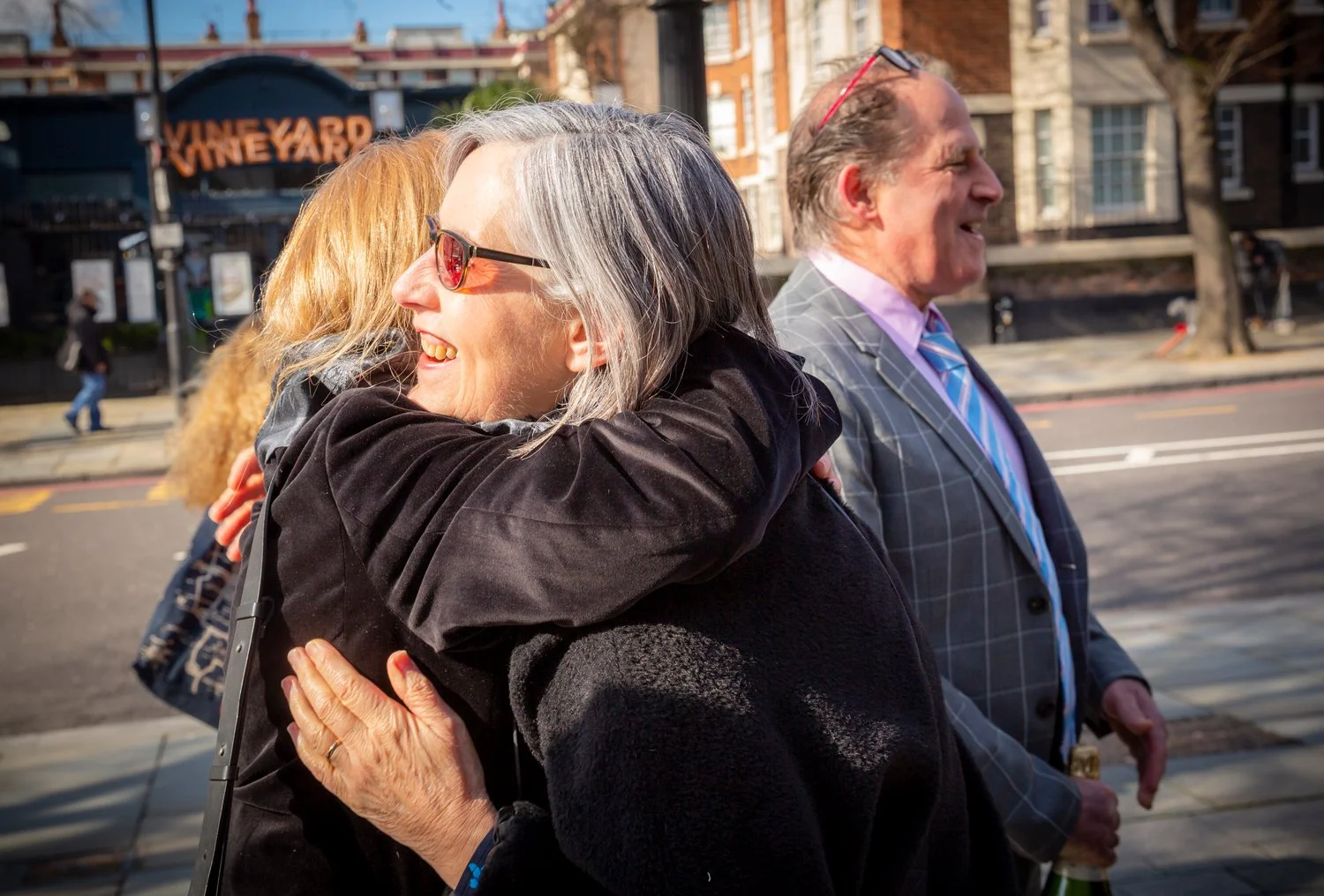 People smiling and hugging outdoors on a city sidewalk, with a sign reading 'Vineyard' in the background.