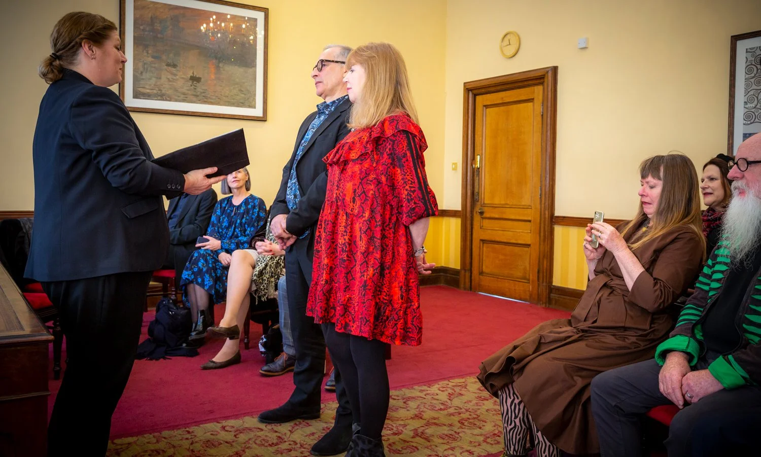 An award or certificate being presented to a woman in a red dress during a formal ceremony in a room with yellow walls and a red carpet. Several people are seated, including a woman in a brown dress taking a photo, a man with glasses, and others watc