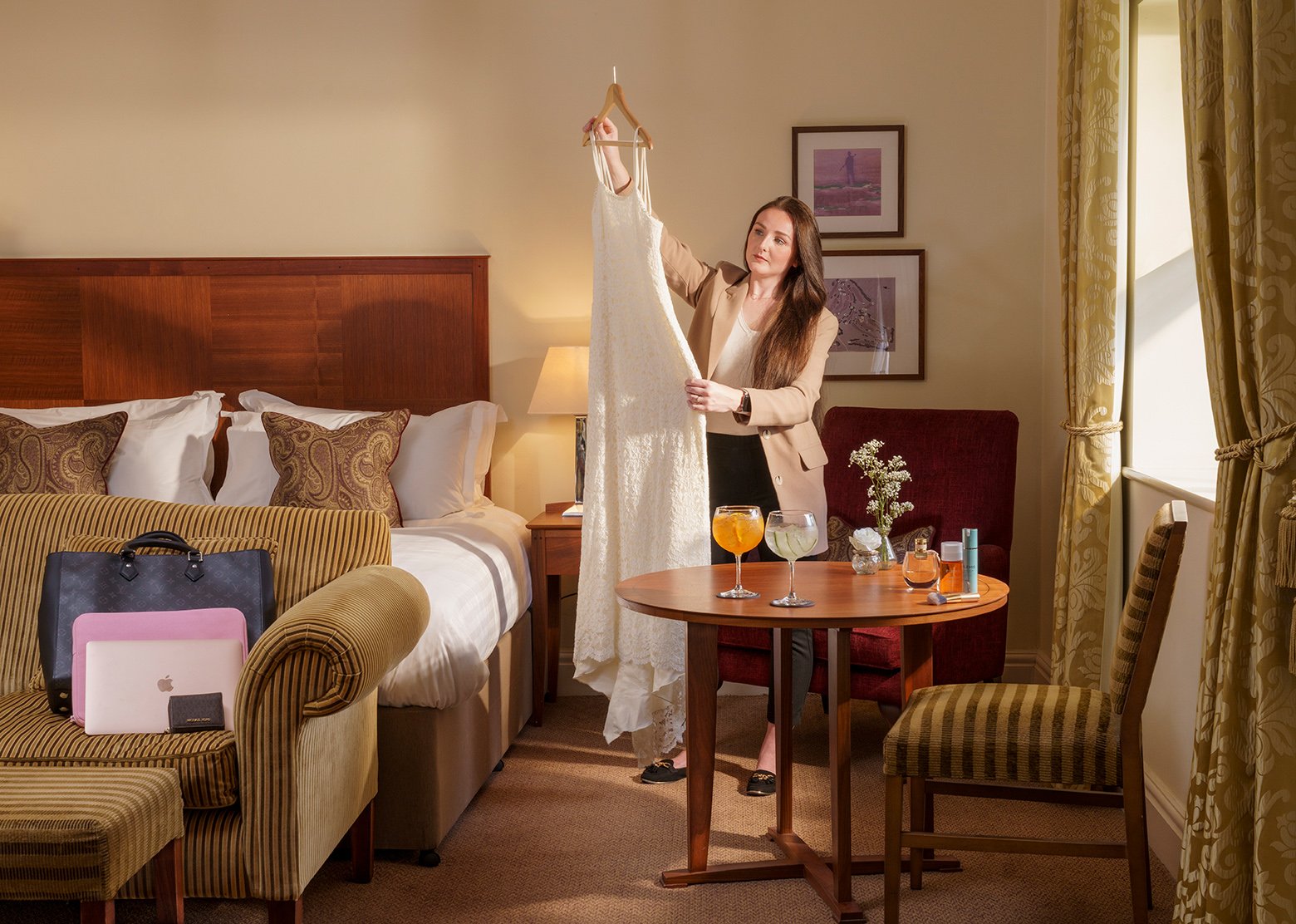 Woman in a beige blazer looking at a white wedding dress while standing in a hotel room with a bed, table with drinks, and luggage.