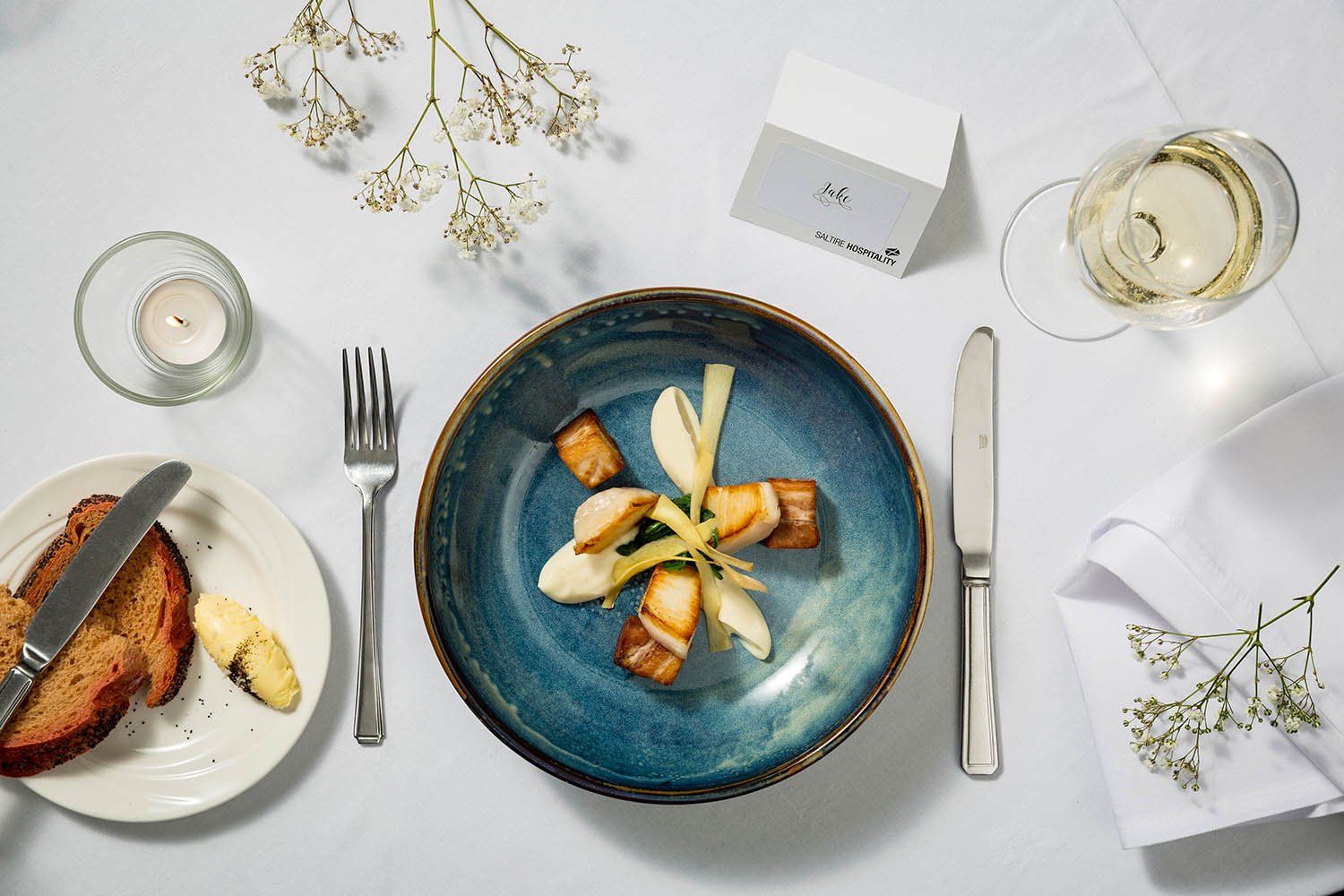 Overhead view of an elegant table setting with a white tablecloth, a blue ceramic plate with small portions of food, a glass of white wine, a lit candle in a glass holder, and some white flowers with green stems.