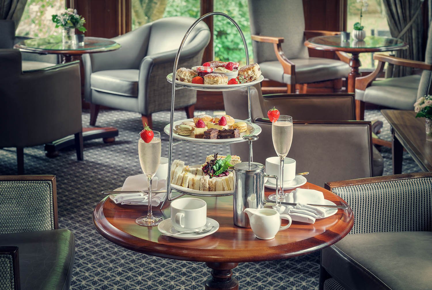 Elegant afternoon tea setup with tiered tray of pastries and sweets, champagne glasses with strawberries, tea cups, and a small pot of cream or milk on a polished wooden table, in a cozy lounge with armchairs and large windows.