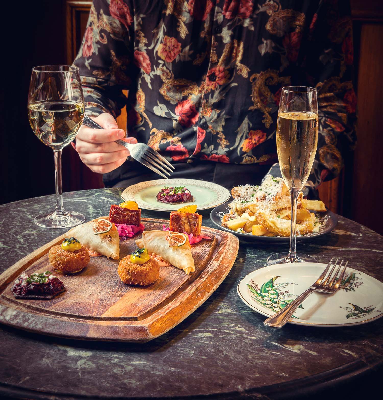 A person in a floral shirt dining at a table with various dishes and two glasses of white wine or champagne.