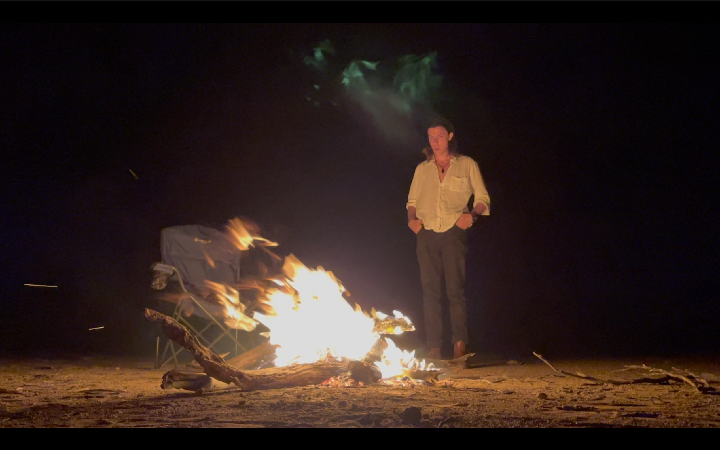A person standing on a beach at night near a campfire with a folding chair nearby. The person has long hair, wears a light-colored shirt and dark pants, with hands in pockets. The night sky is dark and there is smoke rising from the fire.