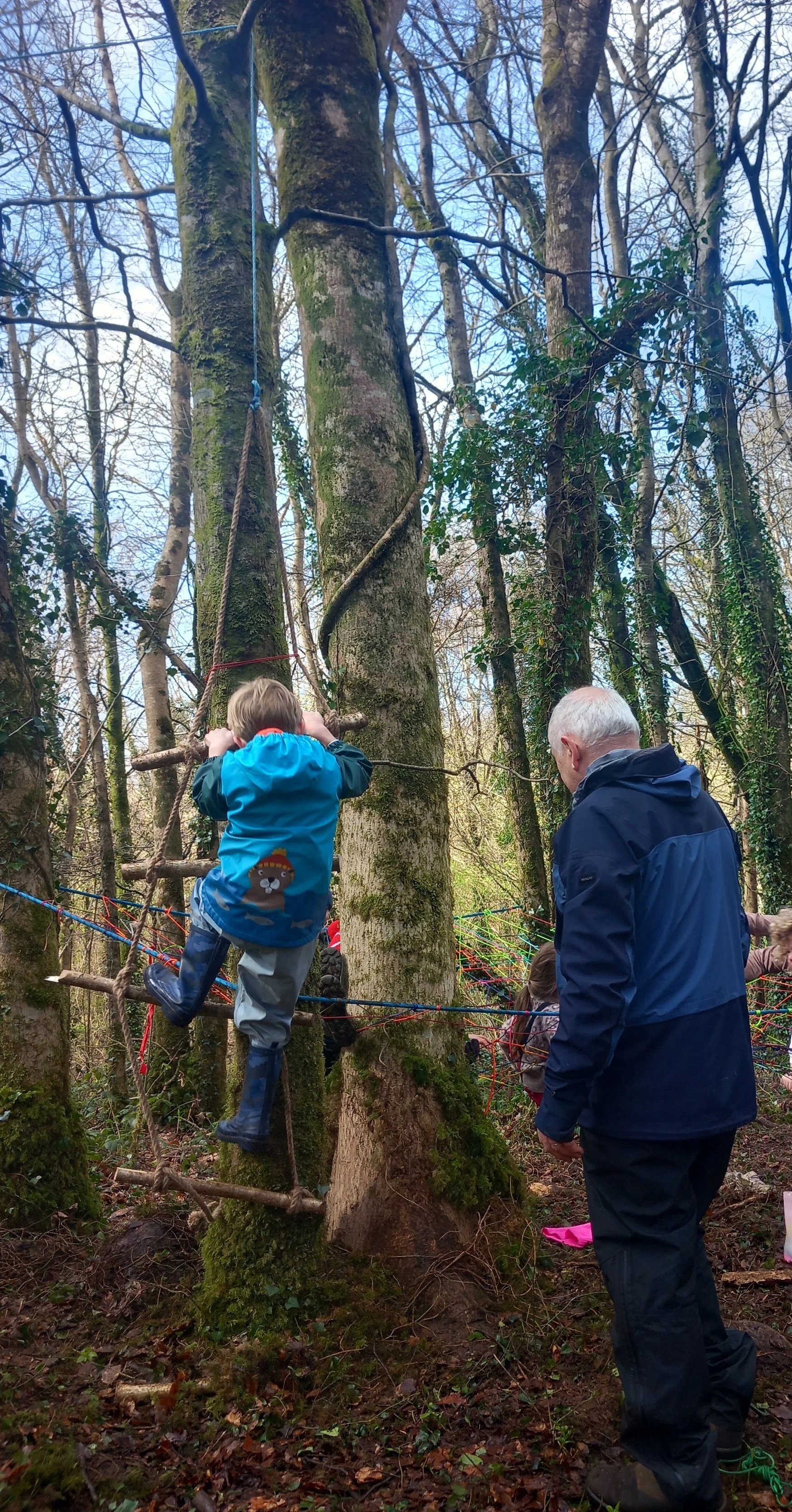 Child climbing a rope ladder in a wooded outdoor area, assisted by an adult man, with other children visible in the background.