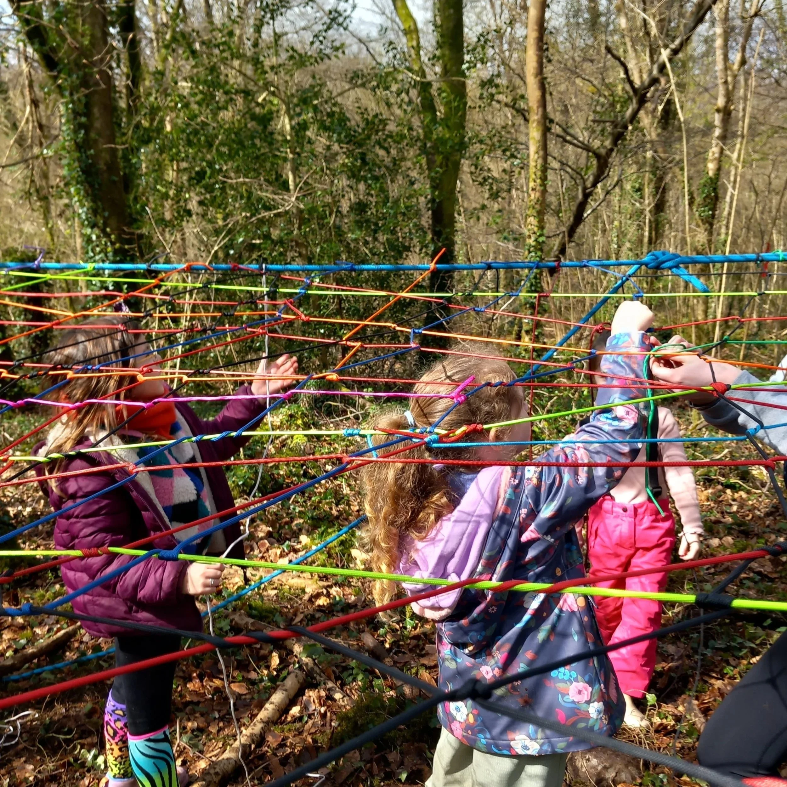 Children playing on a colorful outdoor web structure in a wooded park area.