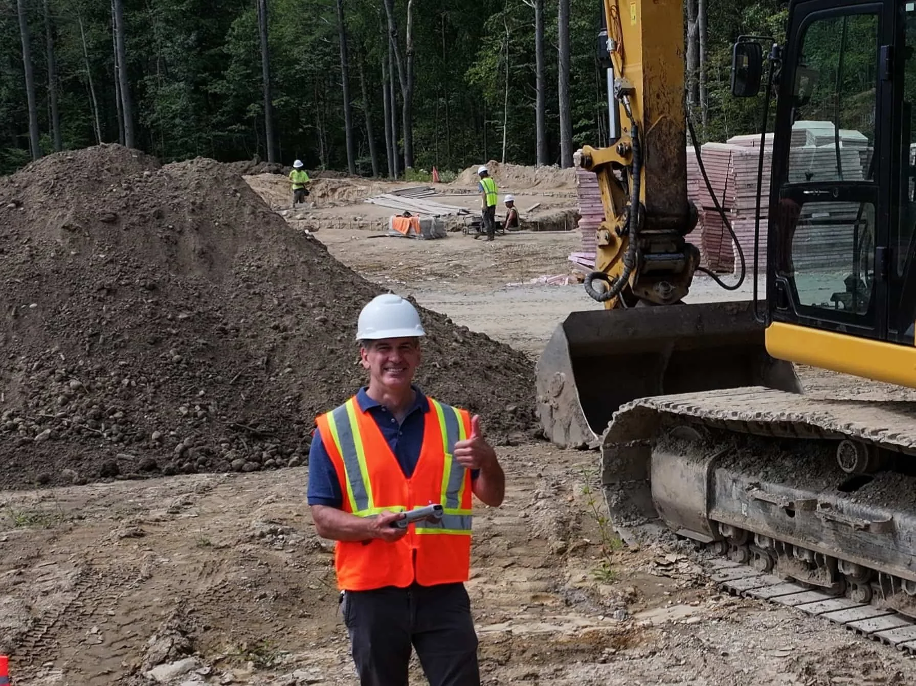 Brian Betts is on-site at a client's construction site standing next to an excavator and a stockpile with construction workers in the background