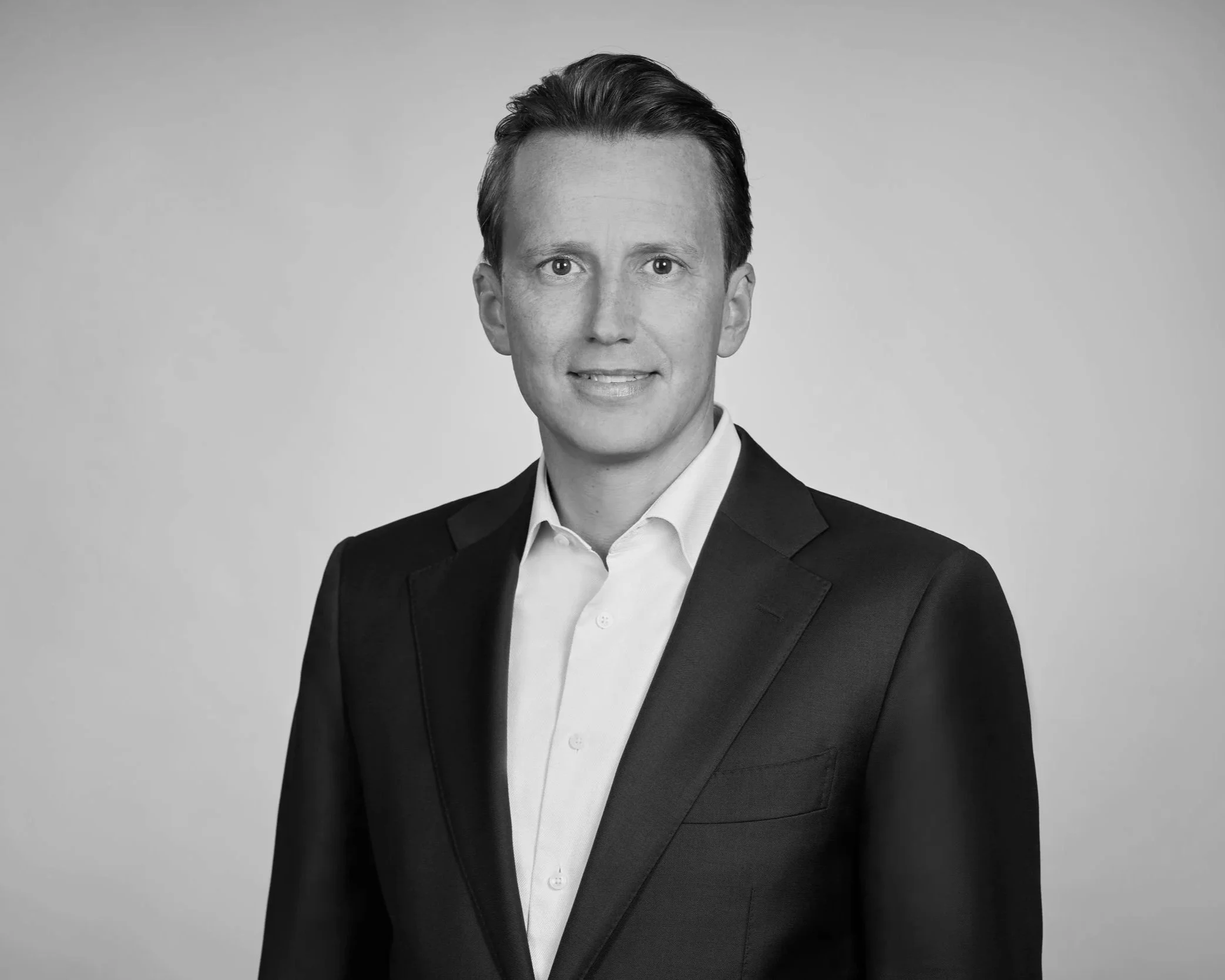 Schwarz-Weiß-Porträt eines Mannes in formeller Kleidung, dunkler Anzug und weißes Hemd, vor einfarbigem Hintergrund.